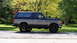 1987 Ford Bronco - A two-tone blue and gray 1987 Ford Bronco is shown from the side on a gravel surface with trees in the background.