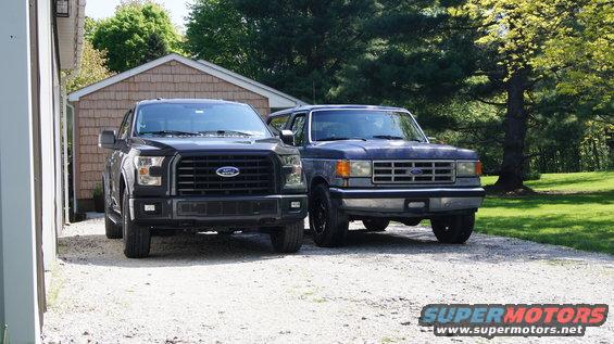 Two Ford vehicles parked on gravel. A blue 1987 Ford Bronco is on the right, and a black Ford F-150 is on the left