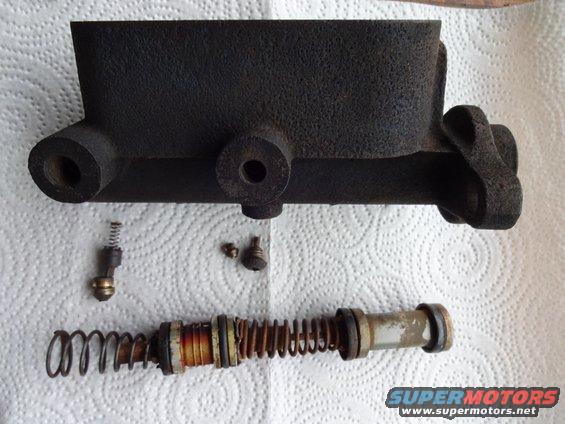 Disassembled brake master cylinder and internal parts laid out on paper towels. Includes cylinder body, piston, spring, and small hardware