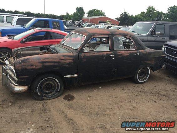 Old rusty sedan, hood up, flat tire, parked in a lot with other vehicles