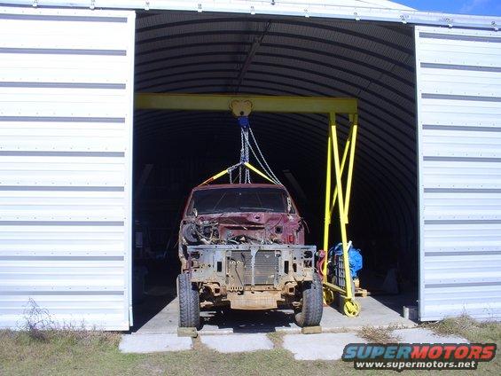 1992 Ford Bronco body, stripped down, suspended by a yellow gantry crane inside a metal building