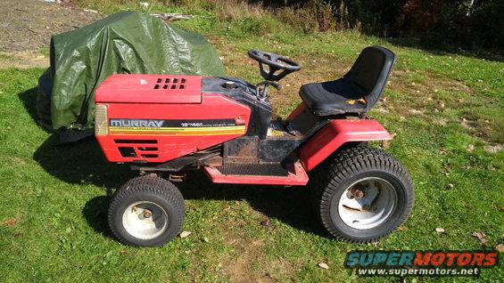Red Murray riding lawn mower with a green tarp covering part of it. The mower has a black seat and steering wheel