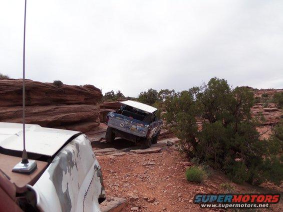 Blue off-road SUV with white roof panel, rear view, on rocky trail. Another vehicle in foreground