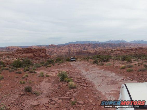 Two off-road vehicles on a dirt track in a desert with red rock formations and mountains in the background. "SUPERMOTORS.NET" logo visible