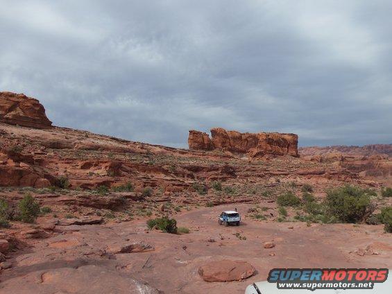 Two off-road vehicles drive on a dirt road through a rocky, desert landscape with red rock formations under a cloudy sky