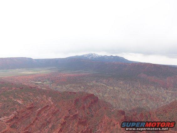 Red rock canyon landscape with distant snow-capped mountains under a cloudy sky. Supermotors.net logo in lower right