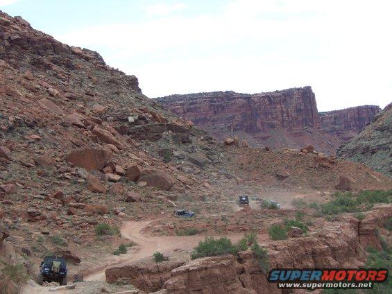 Off-road vehicles navigate a dusty trail through a rocky canyon landscape