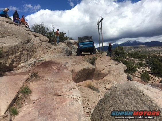Blue SUV on a rocky incline, rear view. People sit on rocks to the left