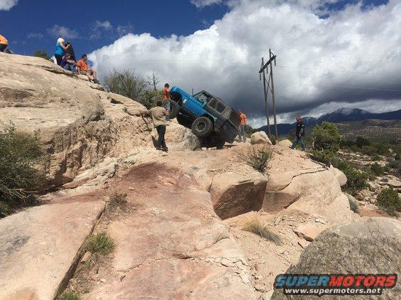Blue SUV on a steep, rocky incline with people watching. Power lines visible in background