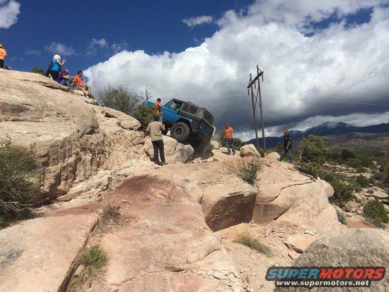 Blue SUV attempting to climb a steep, rocky incline with people watching from above