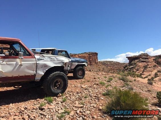 Two battered SUVs parked on a rocky desert trail, one red and white, the other blue and white