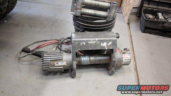 worn electric winch with red and black cables, sitting on concrete floor next to a bucket with rollers