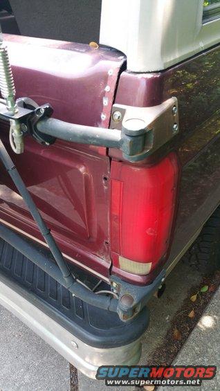 Rear view of maroon SUV showing tailgate hinge, bumper, and taillight. A spare tire carrier is mounted to the tailgate
