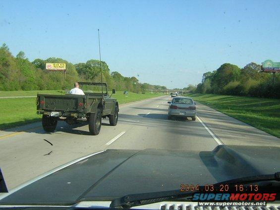 Green military-style jeep driving on highway, rear view. Driver visible