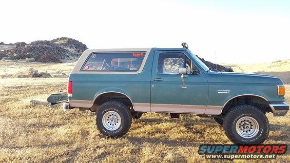 Green Ford Bronco parked in a dry, grassy field with rocky hills in the background. It has a tan top and off-road tires