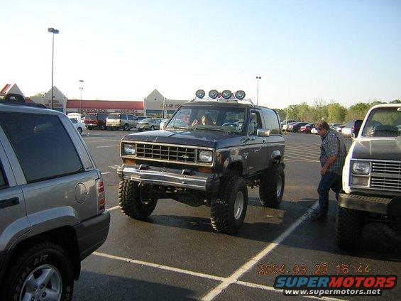 Lifted dark blue Ford Bronco II with roof lights parked in a lot next to a silver SUV and a white pickup
