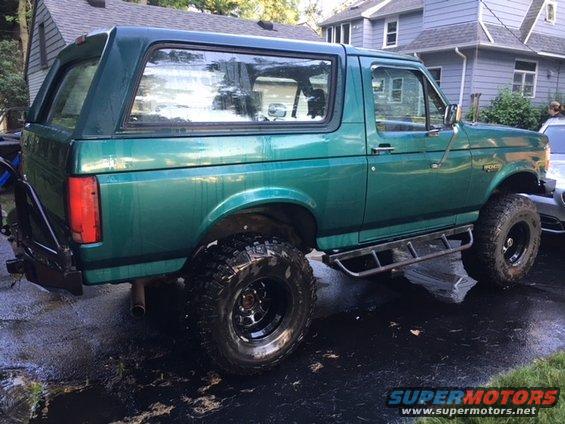 Green 1996 Ford Bronco with aftermarket wheels and side steps parked on wet pavement
