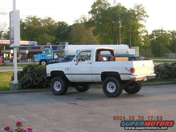 White Chevrolet pickup truck with 4x4 decal on the side, parked at a gas station. Blue truck and tanker trailer in background
