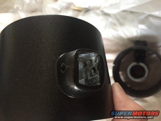 Black cylindrical part with a square opening, held by a hand. Another part is visible in the background
