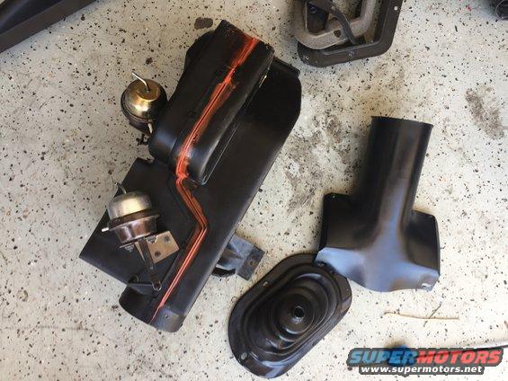 Black car parts including a heater core housing with orange accents and vacuum actuators, a shifter boot, and ducting, laid out on a concrete floor