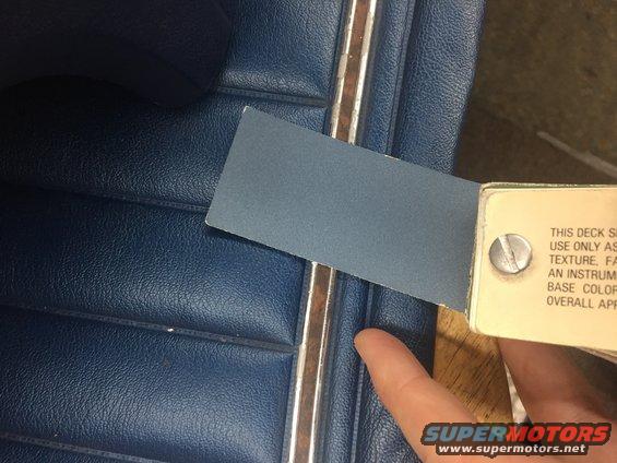 Blue vinyl seat with chrome trim and a swatch of blue material held by a finger. Text is visible on a swatch holder