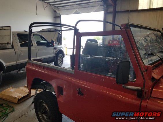 Red Land Rover Defender with roll cage, side view. A silver pickup truck is parked behind it