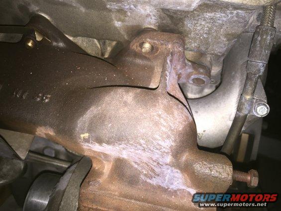 Rusty exhaust manifold with visible cracks and white residue. A bolt is attached to a bracket