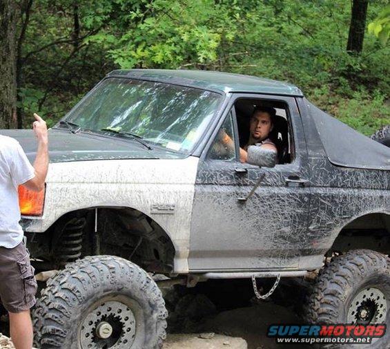Man driving a lifted Ford Bronco with large tires on a trail. Another person is outside the vehicle pointing