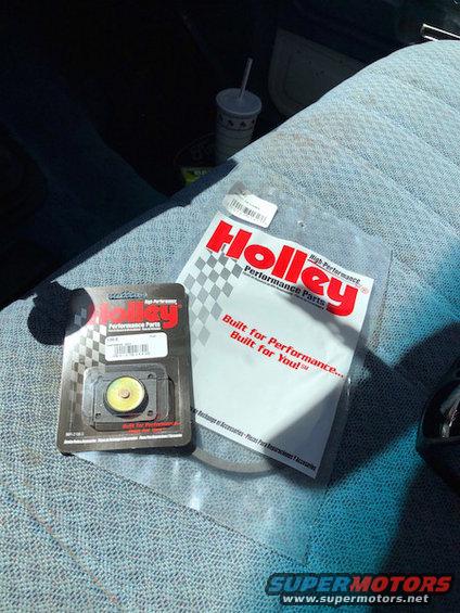 Holley performance parts packaging and a small yellow part on a blue patterned car seat next to a cup with a straw
