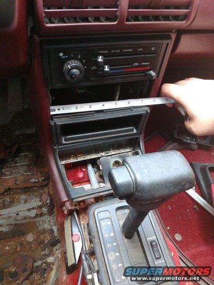 Interior of a car with a measuring tape across the center console, showing the gear shifter and radio area. The floor shows rust and damage