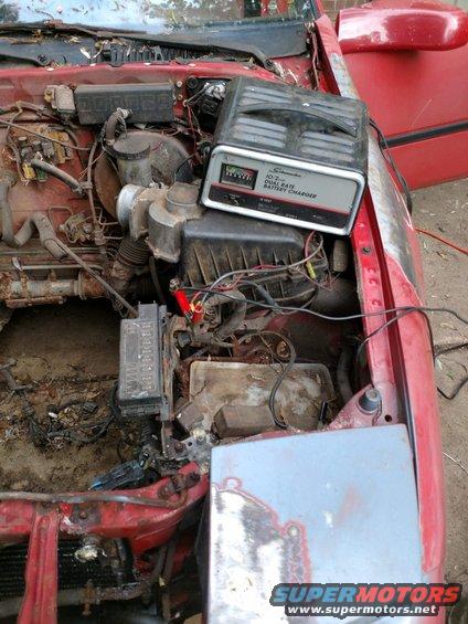 Red car engine bay with battery charger on top, showing rust and exposed wires