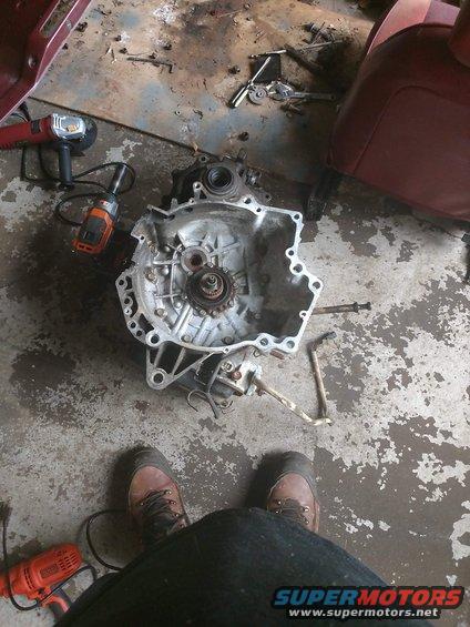 Disassembled transmission on concrete floor with tools and a red seat. Transmission is silver and appears to be undergoing repair