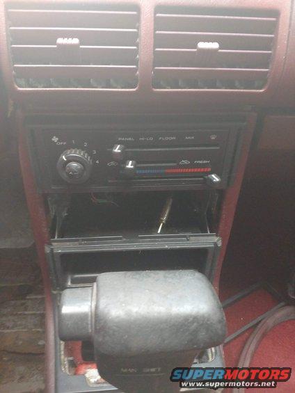 Car interior dashboard with maroon trim, showing air vents, radio controls, and gear shifter. A tool is in the open storage compartment