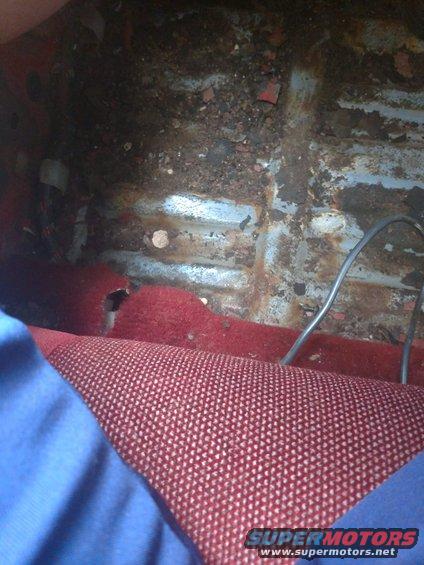 Car interior showing heavily rusted floor with torn red carpet and exposed metal
