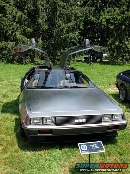 DeLorean DMC-12 with gullwing doors open, parked on grass. Sign says 1981 DeLorean DMC-12