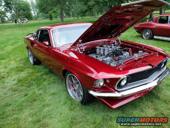 Red 1969 Ford Mustang Mach 1 with engine exposed, parked on grass next to a red classic car