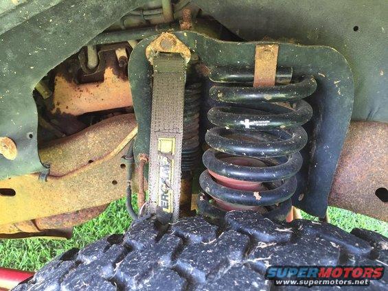 Close-up of a vehicle's front suspension with a coil spring, shock absorber, and tire tread. A yellow strap is visible