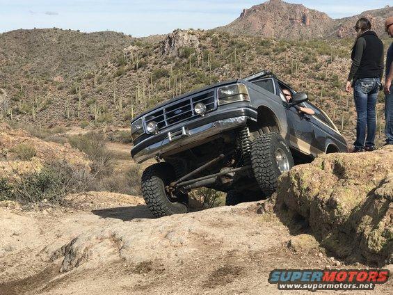 Ford Bronco climbing rocks on a desert trail, driver visible, two people watching