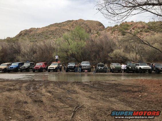 Multiple SUVs and off-road vehicles gathered in a muddy, shallow stream with a desert mountain in the background
