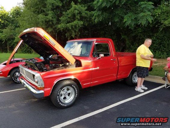 Red 1970s pickup truck with hood open, parked next to a red sports car. A man stands beside the truck