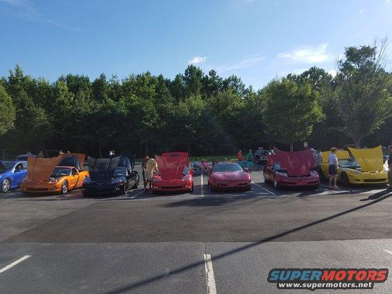 Several sports cars with hoods open parked in a lot with trees and a blue sky