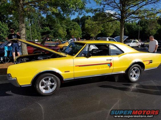 Yellow coupe with red and blue racing stripes on the side, chrome wheels, hood open. Other cars visible in background