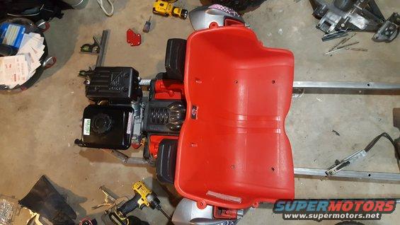 Go-kart frame with a black engine and red plastic seat. Tools and parts are scattered around