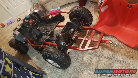 Toy ATV flipped over, showing its underside and internal wiring. Red plastic seat is detached
