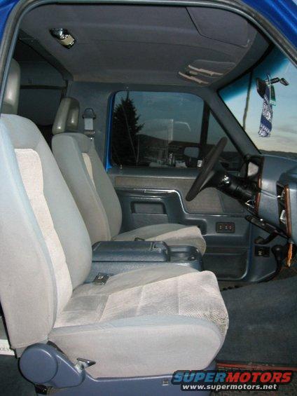 Interior of a blue truck with cloth seats, center console, and steering wheel. Outside view shows trees and sky