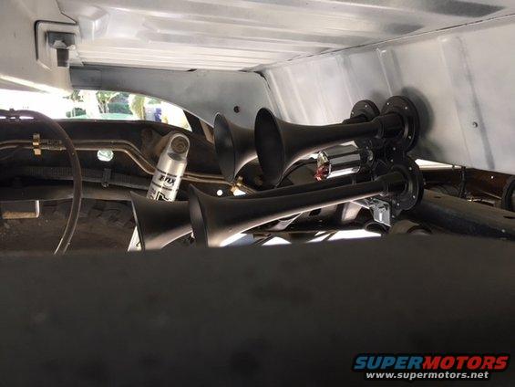 Four black air horns mounted on a bracket under a vehicle's chassis, with a silver shock absorber visible