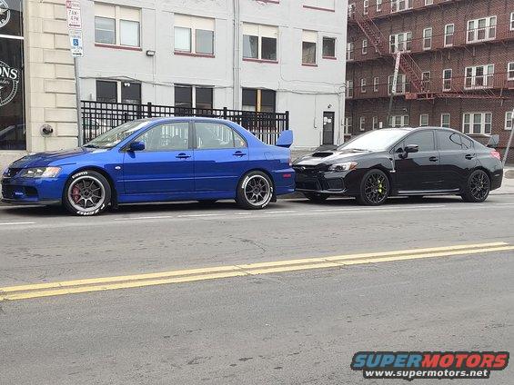 Blue Mitsubishi Lancer Evolution IX sedan with a rear spoiler, parked on the street next to a black Subaru WRX