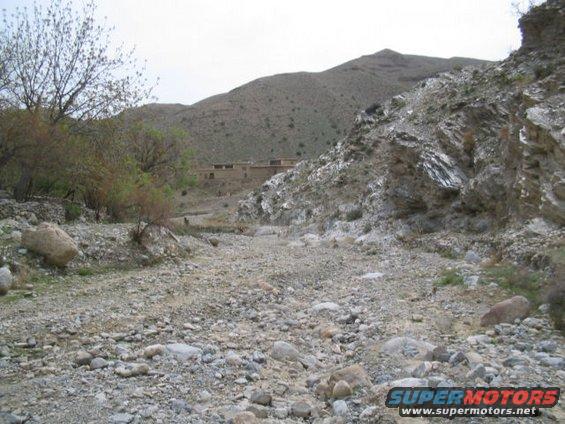 Dry, rocky riverbed leading to buildings in a mountainous area. Overcast sky
