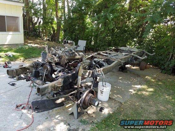 Disassembled vehicle chassis on concrete with engine and suspension parts scattered around. A white bucket and tools are visible