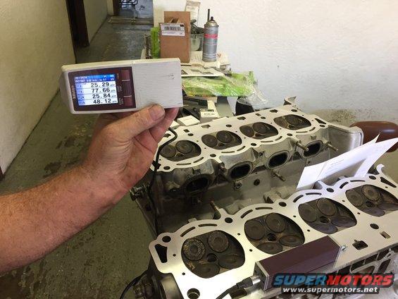 Hand holding digital measurement tool over engine cylinder head. Tool shows readings like 25.29 and 77.66